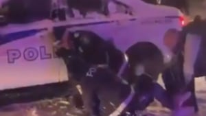 Deux agents tentent d'immobiliser un jeune homme au sol, devant un véhicule de police.