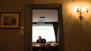 Un homme, assis à son bureau, est aperçu à travers le cadre de la porte dans le corridor de l'hôtel de ville.