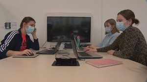 Trois jeunes femmes assises à une table, chacune devant un ordinateur portable. Elles regardent l'écran et portent toutes un masque.