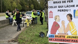 Une pancarte arbore une caricature qui dépeint le président Macron et un gilet jaune en séance de tir au poignet. « Celui qui lâche a perdu. Les gilets jaunes ne lâcheront pas. ». En arrière-plan, des manifestants réunis à côté d'une route.
