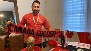 Un homme portant un gilet de l'équipe canadienne de soccer, tenant un foulard rouge où est inscrit «Canada soccer». Divers articles représentant l'équipe se trouvent devant lui sur une table.