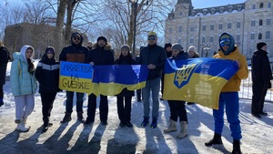 Un groupe de personne devant l'Assemblée nationale. Certains tiennent devant eux un drapeu ukrainien déplié. Le ciel est bleu et il y a de la neige au sol.
