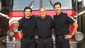 Un père et ses deux fils se tiennent bras dessus, bras dessous en uniforme de pompier devant leur camion. 