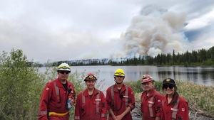 Trois hommes et deux femme pompier posent au bord d'un lac avec de la fumée qui sort de la forêt derrière eux. 