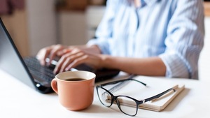 Une femme travaille sur un ordinateur portable. Une tasse de café et des lunettes se trouvent en avant-plan.