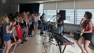 Deux femmes chantent et jouent de la guitare devant un groupe d'une vingtaine de jeunes filles. 