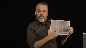 L'homme tient une photo en noir et blanc où l'on voit plusieurs personnes et il en montre une du doigt.