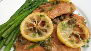 Photo of Italian recipe called chicken piccata.  This is chicken scaloppine or chicken breast cutlets cooked with a sauce made of capers, white wine, lemon slices and butter.  Parsley is sprinkled on top.  Asparagus is served with the chicken as the vegetable.