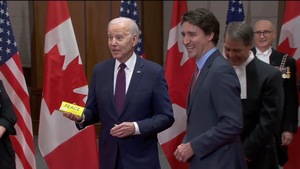 Joe Biden pose à côté de Justin Trudeau devant des drapeaux canadiens et américains. Il tient dans sa main une boîte où on peut lire le mot Peace.
