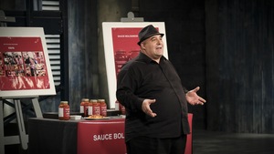 Il est devant un présentoir présentant ses sauces tomates dans des bocaux et il porte un chapeau.