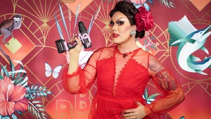 La drag queen habillée de rouge tient une perceuse dans la main. Elle pose devant le mur colorée de l'émission.