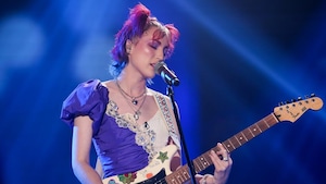 Une femme qui chante et joue de la guitare. Elle porte une robe violette et blanche. 