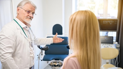 Un médecin aux cheveux et à la barbe blanche est devant une jeune femme de dos et pointe une chaise d'examen.