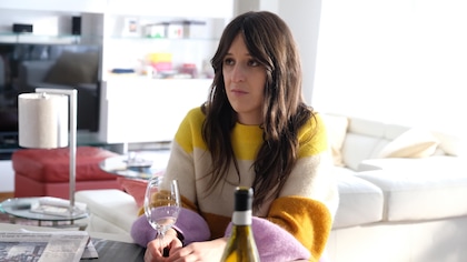 Une jeune femme est assise à table. Elle tient un verre de vin blanc.