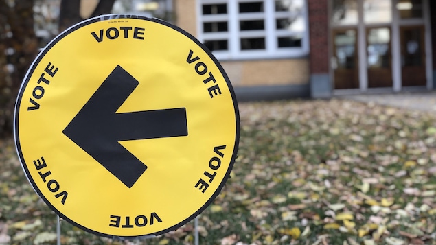 Une pancarte sur une pelouse parsemée de feuilles. La pancarte est ronde. On y voit une flêche au centre pointant vers la gauche. Le mot vote est écrit six fois en cercle.