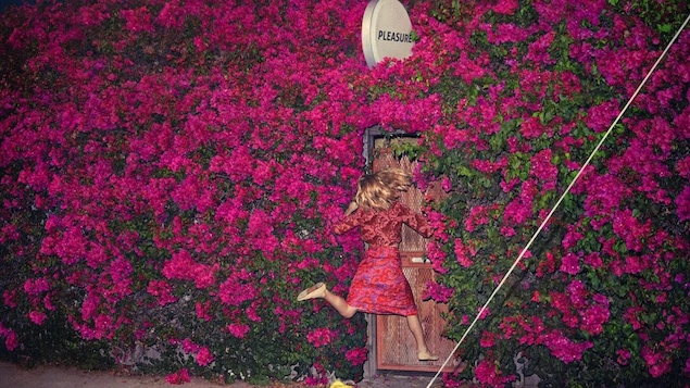 A woman jumps in front of a wooden door that's set in a large wall of pink rhododendron bushes. A circular sign above the door has one word on it, "Pleasure."