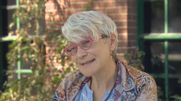 Gros plan sur Francine Proulx-Kenzle, elle porte une chemise bleue avec un foulard à motifs. Elle a les cheveux courts et porte des lunettes.