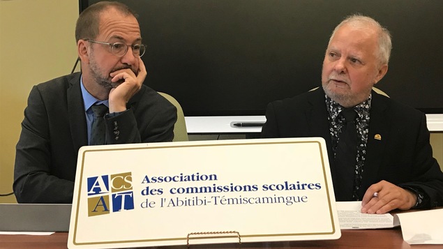 Deux hommes parlent lors d'une conférence de presse derrière une affiche de l'Association des commissions scolaires de l'Abitibi-Témiscamingue.