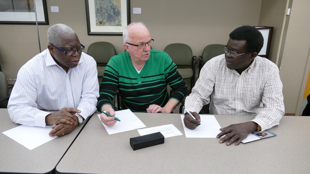 Trois hommes assis à une table discutent en tenant devant eux leurs stylos sur des feuilles de papier. Trois hommes assis à une table discutent en tenant devant eux leurs stylos sur des feuilles de papier.