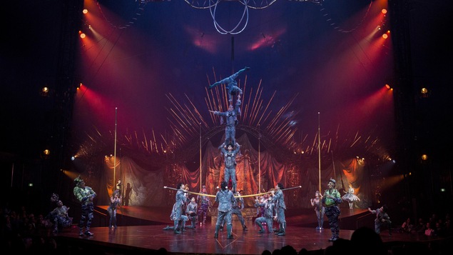 Trois acrobates costumés se tiennent en équilibre sur une pôle tenue par d'autres artistes du Cirque du soleil, pendant que d'autres personnages costumés les observent sur la scène.