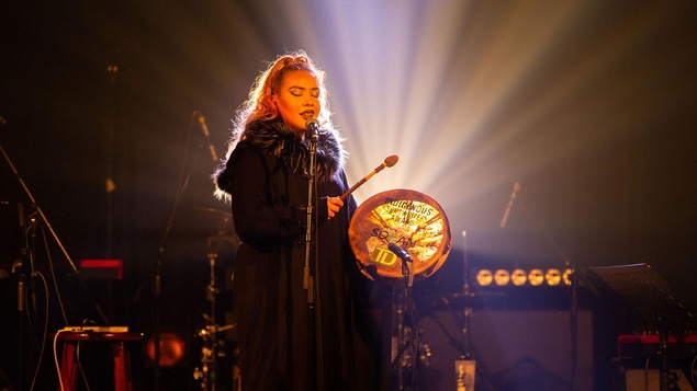 La chanteuse chante sur scène devant un micro et joue du tambour.