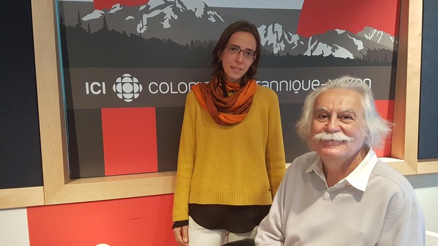Un homme avec une moustache blanche et des cheveux mi-longs blancs assis dans une chaise et derrière lui debout une femme petite qui porte un pull jaune et des lunettes.