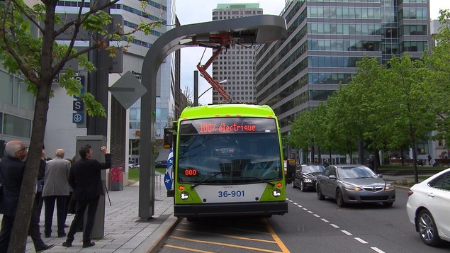 Un autobus qui affiche sur son écran frontal « 100 % électrique » est arrêté le long d'un trottoir. Un bras mécanique sort de son toit et est accroché à un poteau installé en bordure de trottoir.