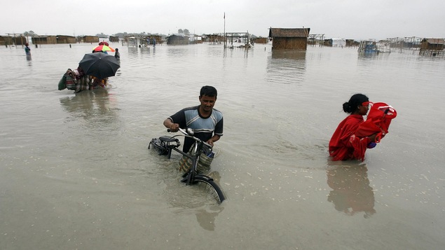 Une inondation détruit un village. Un homme pousse son vélo dans l’eau jusqu’à la taille. Une femme transporte ses biens.