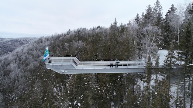 On voit un belvédère, en pleine nature, positionné au-dessus d'une falaise. Il est entouré d'arbres, de conifères principalement. C'est l'hiver, alors les branches des arbres sont parsemées de neige. Deux personnes se trouvent sur le belvédère. Au bout du belvédère, il y a un drapeau.