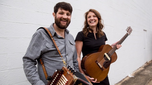 Un homme avec un accordéon et une femme tenant une guitare rient dans une ruelle devant un mur de briques blanc.
