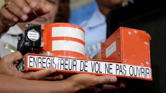 Des personnes tiennent un enregistreur de vol dans leurs mains. L'appareil est une sorte de boîte en métal de couleur orange vif.