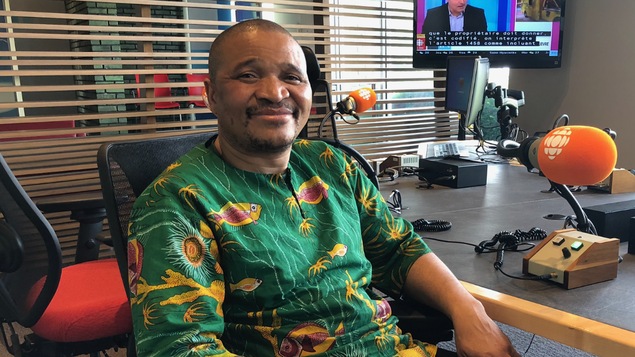 Portrait d'un homme souriant. Il est attablé dans un studio de radio.
