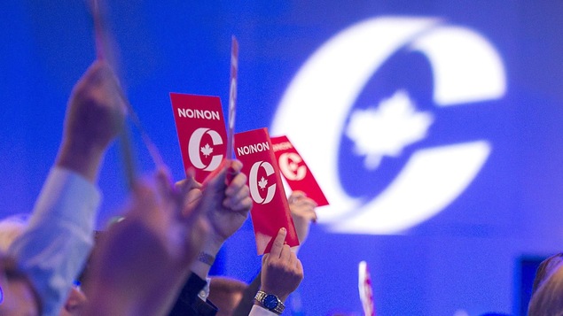 Des personnes assise dans une grande salle, vues de profil. votent en tenant des cartons en l'air.