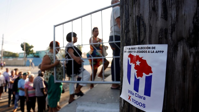 Des hommes et des femmes sont photographiés en file, lors d'une journée ensoleillée. Ils sont légèrement flous, puisque le focus de la caméra est effectué sur une petite affiche en avant-plan qui dit, en espagnol, « 11 mars, élection des députés et des délégués à la APPP ». 