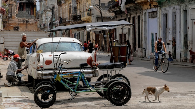 Une voiture et un vélo-taxi sont garés en bordure de la rue, dans un quartier pauvre de La Havane.