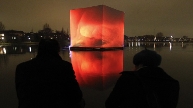 Deux personnes regardent une étendue d'eau sur laquelle flotte un énorme cube illuminé.