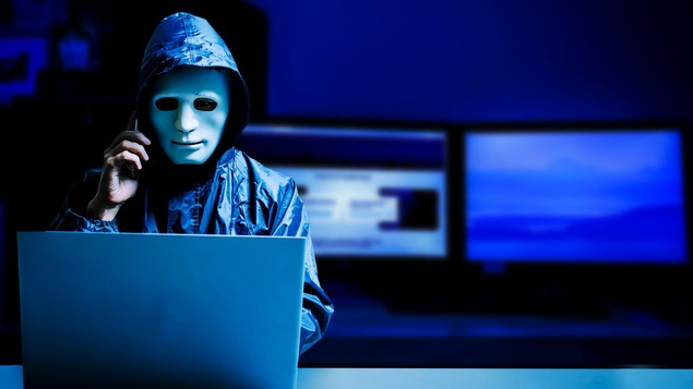 Un pirate informatique portant un masque parle au téléphone devant un ordinateur portable.