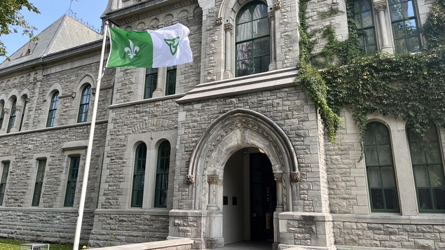 Un drapeau vert et blanc flotte devant un édifice aux briques grises.