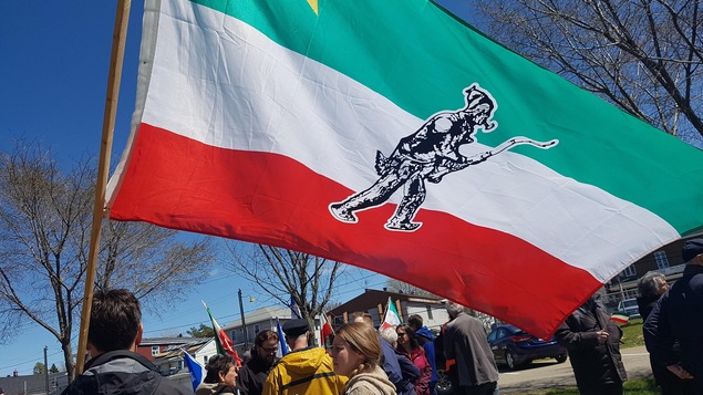 Une dizaine de personnes sont réunies dans un parc. À l'avant-plan, un homme tient un grand drapeau aux couleurs des Patriotes.