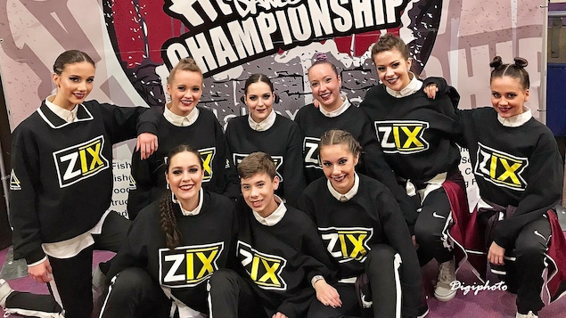 Huit danseuses et un danseur assis sur deux rangs devant l'affiche du championnat national.