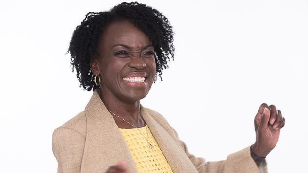 Une femme noire avec une robe jaune et une veste beige qui a les bras levés.