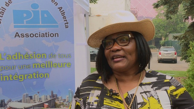 Une femme avec des lunettes de vue et un chapeau de paille, devant une affiche de l'association PIA.