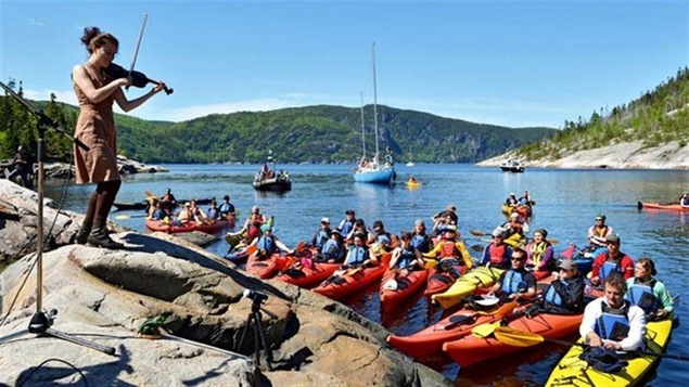 Violonniste sur une pierre devant un lac qui joue devant des kayakistes.