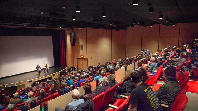 Le public est assis dans la salle de spectacle face à la scène où est assis Harold Arsenault.