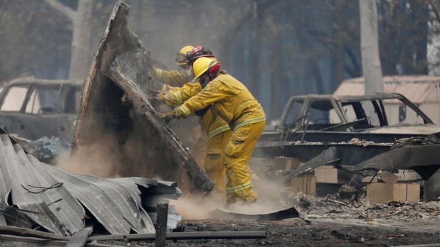 Deux pompiers avec des masques et leurs casques de protection repoussent de lourds débris d'une maison calcinée.