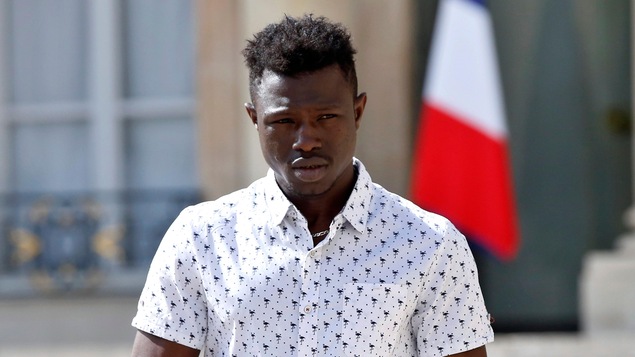 Un jeune Noir, aux cheveux courts et frisés, est photographié de face alors qu'il regarde légèrement vers sa gauche. Il porte une chemise à manches courtes et se trouve devant un drapeau français.