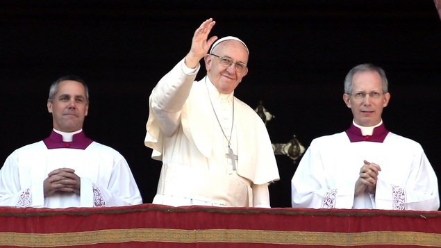 Un homme portant des habits pâle et une calotte pâle lève sa main droite en souriant, comme s'il saluait des personnes. Derrière lui se tient deux autres hommes qui ont les mains croisées et qui sourient légèrement.