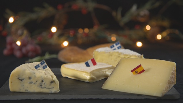 On voit des fromages disposés sur une table. Certains arborent un drapeau québécois, un autre un drapeau français et un autre un drapeau espagnol.