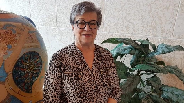 Une femme âgée aux cheveux courts et avec des lunettes rondes regarde l'objectif en souriant.