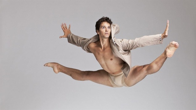 Le danseur Guillaume Côté est posé alors qu'il fait le grand écart dans les airs. On comprend qu'il a jumelé un saut et la figure du grand écart. l'artiste est vêtu d'une culotte et manteau court beige.